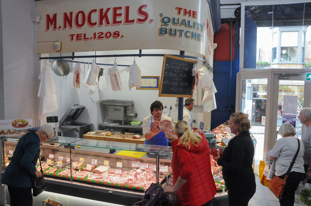 The M. Nockels counter inside Scarborough Market Hall, with the painted fascia sign, hanging aprons and a full glass display counter